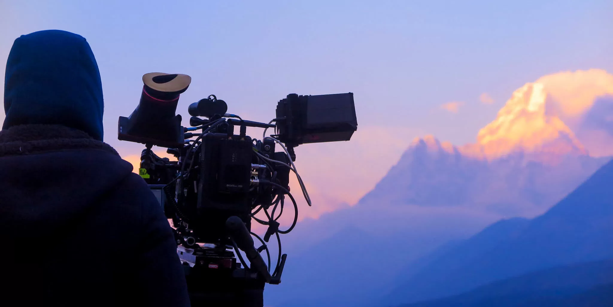 Camera operator in a hooded jacket framing a snow-lit mountain peak at dusk