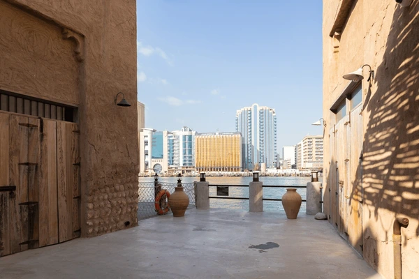 Heritage lane with wooden doors and clay pots opening onto Dubai Creek with modern buildings opposite
